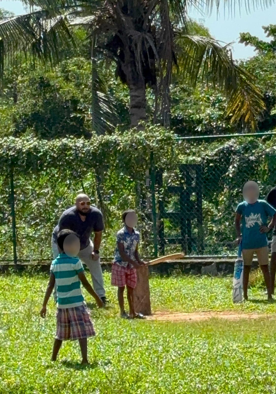 A Coderra team member keeping wicket during a game of cricket with children on the grassy field at the centre, children's faces blurred for privacy.