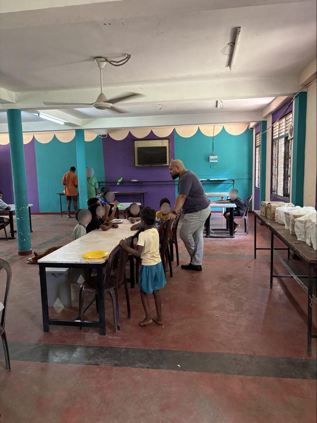 A Coderra team member chatting with children seated at a long table in the centre's brightly painted dining hall, with boxed lunches lined up on a side table.